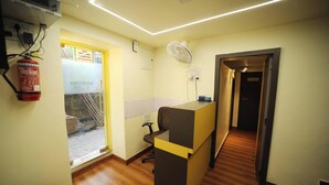A reception area with a wooden floor, a reception desk, a fire extinguisher, a fan, and a door leading to another room. - CABINSTAY MAROL- THE HUBZ (Mumbai)
