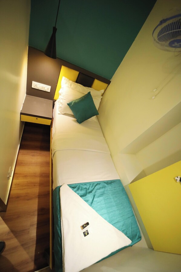 Una habitación de hotel pequeña y estrecha con una cama, una mesita de noche y un ventilador de pared.