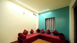 A room with a sectional sofa, wooden flooring, a teal accent wall, and a window with curtains. - CABINSTAY MAROL- THE HUBZ (Mumbai)