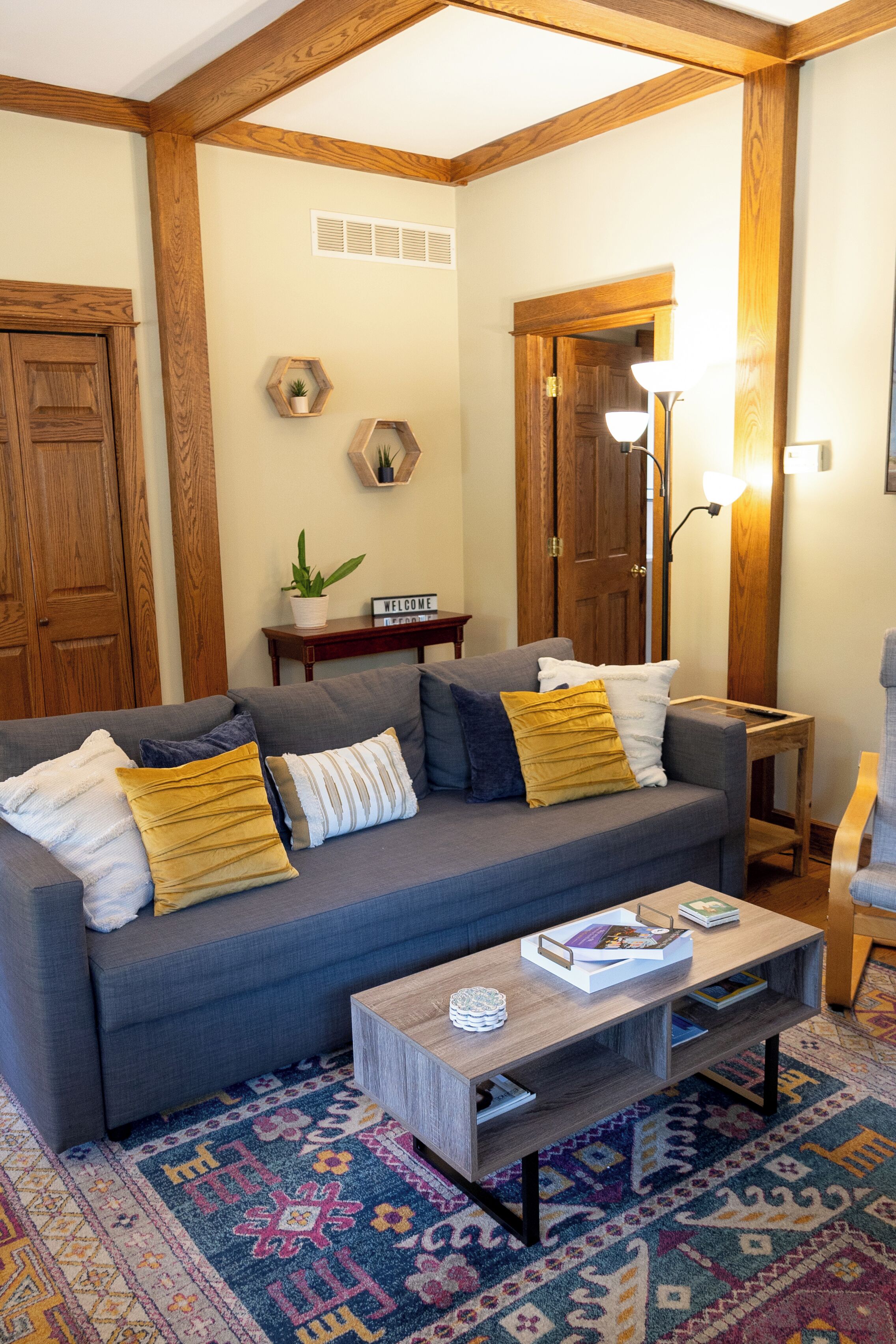 Un salon avec un canapé gris, des poutres en bois, un tapis à motifs et une petite table d’appoint.
