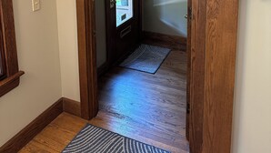 Une porte en bois donne sur un couloir où se trouve un tapis à motifs et une fenêtre offrant une vue sur la verdure.