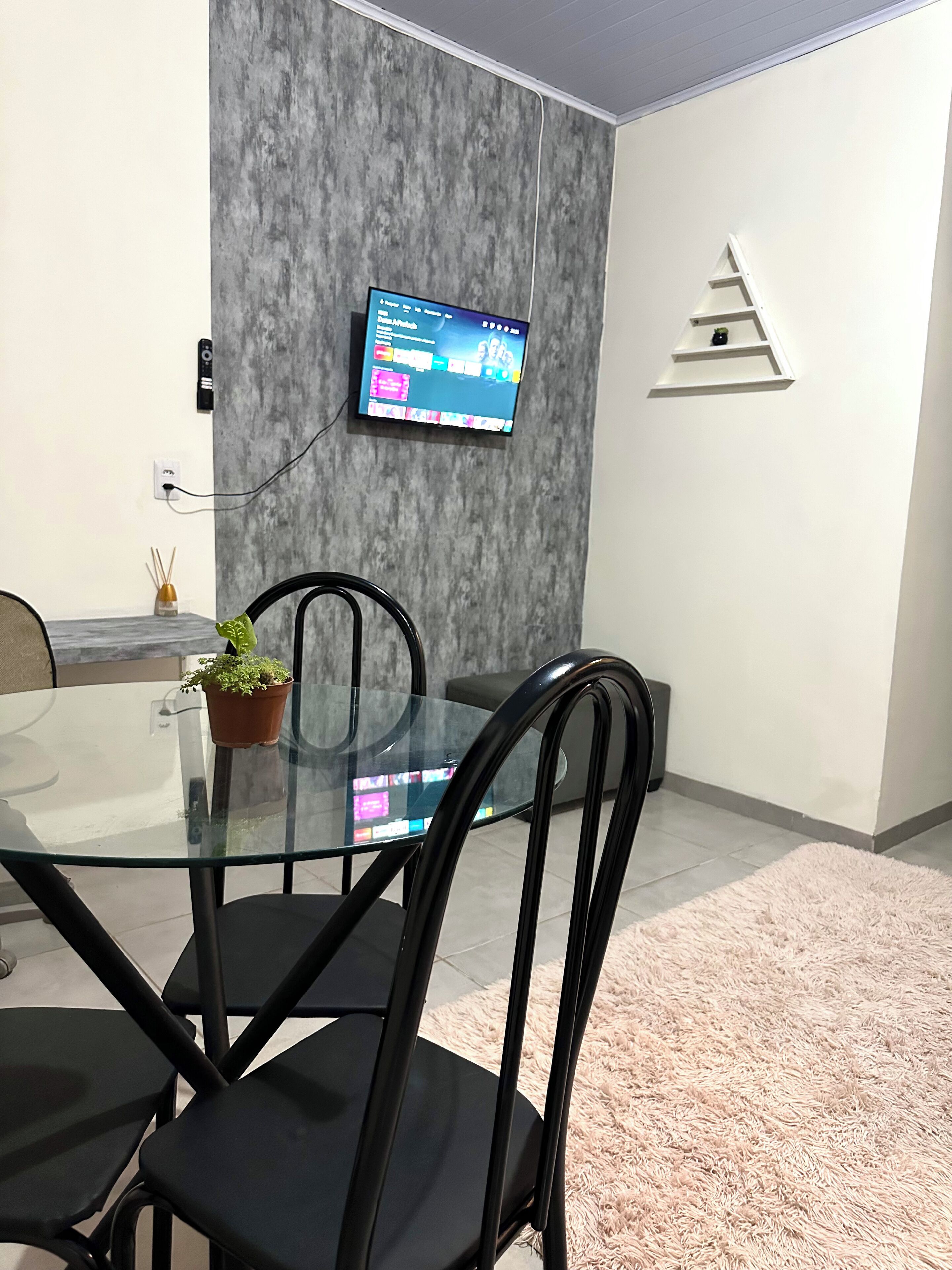 Un comedor moderno con mesa de vidrio, sillas negras, televisor montado y pared gris con textura.