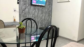 Un comedor moderno con mesa de vidrio, sillas negras, televisor montado y pared gris con textura.