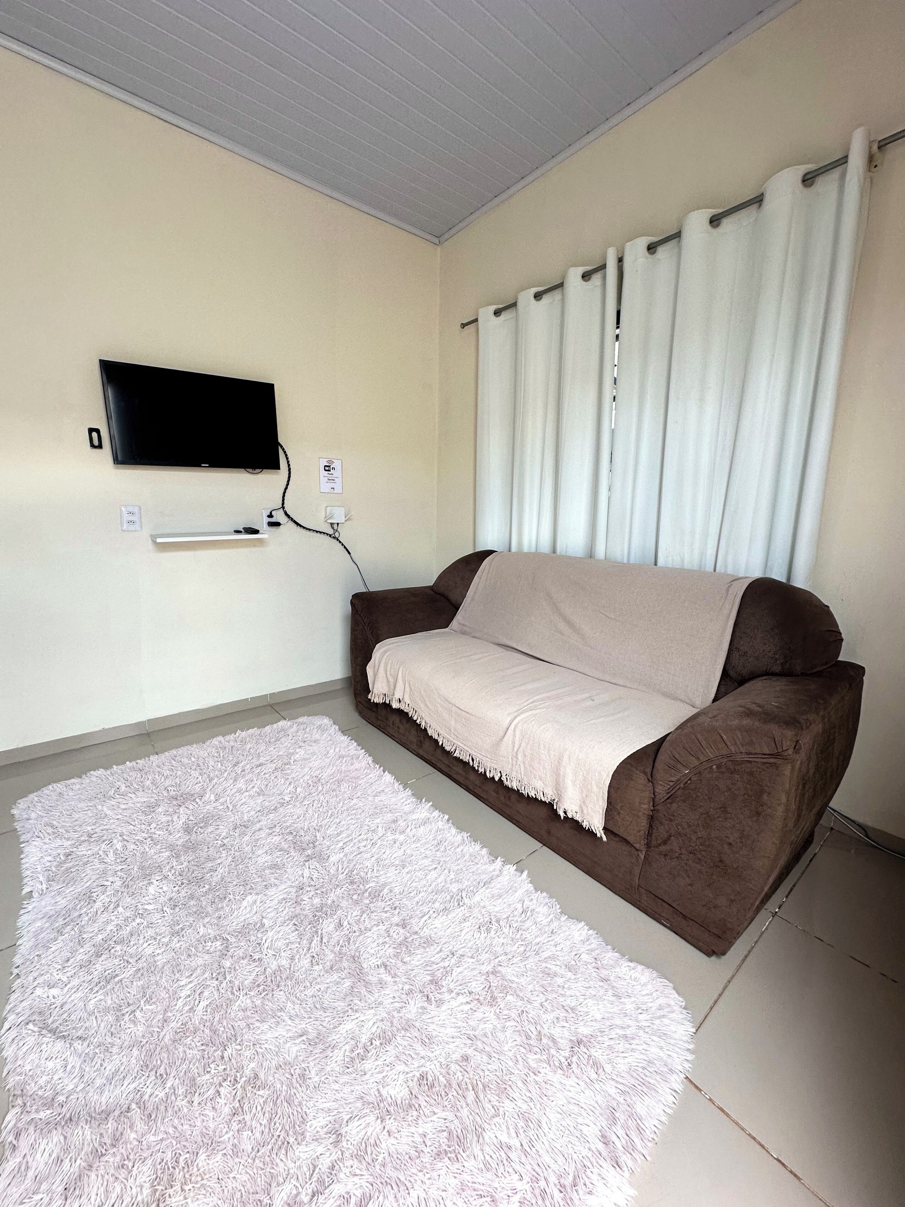 Uma sala de estar com televisão de tela plana na parede, um sofá marrom com manta branca e um tapete felpudo branco no chão.