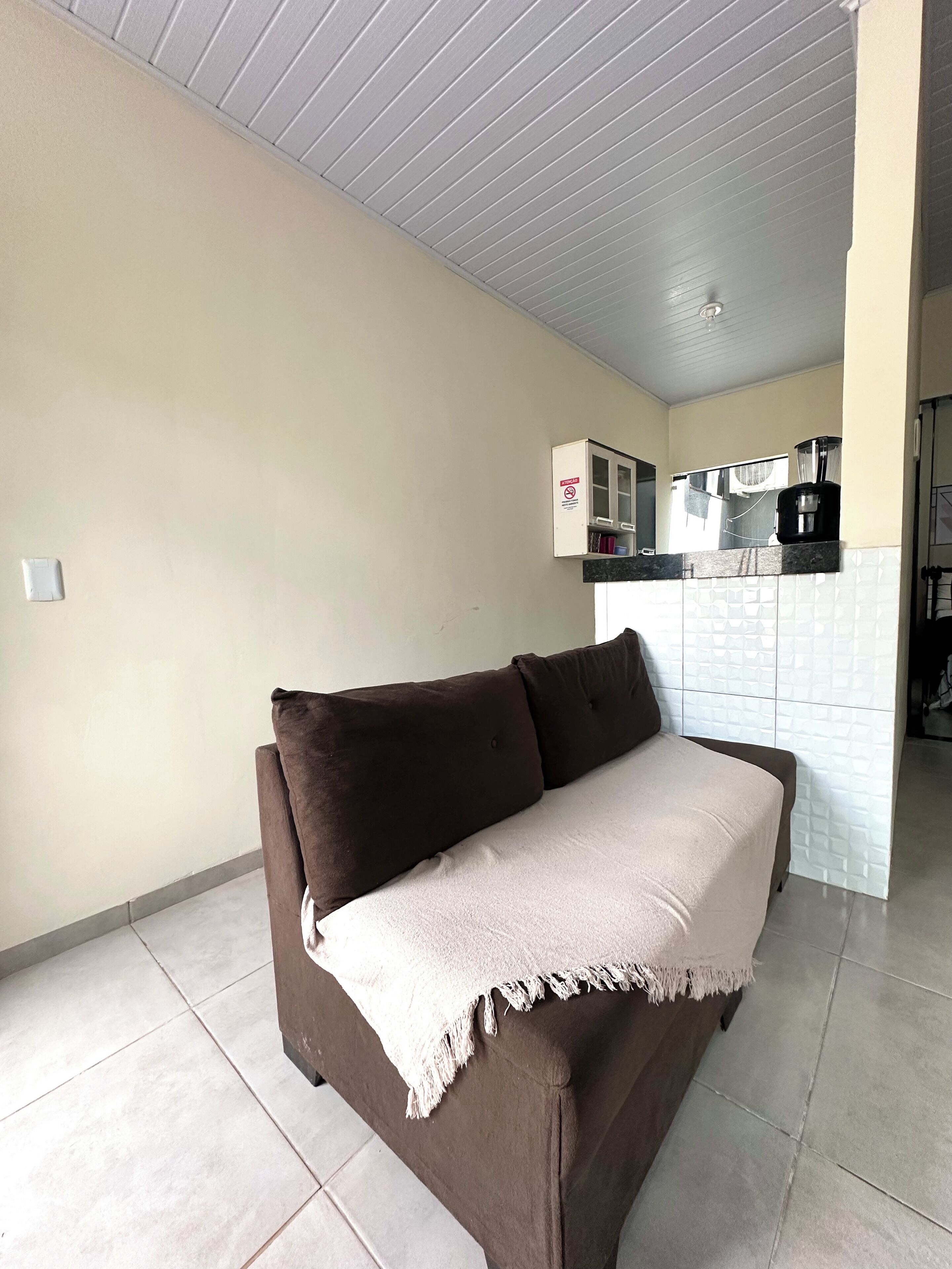 A living room with a brown sofa and a white throw blanket. There is a kitchen area in the background with a coffee machine and cabinets.