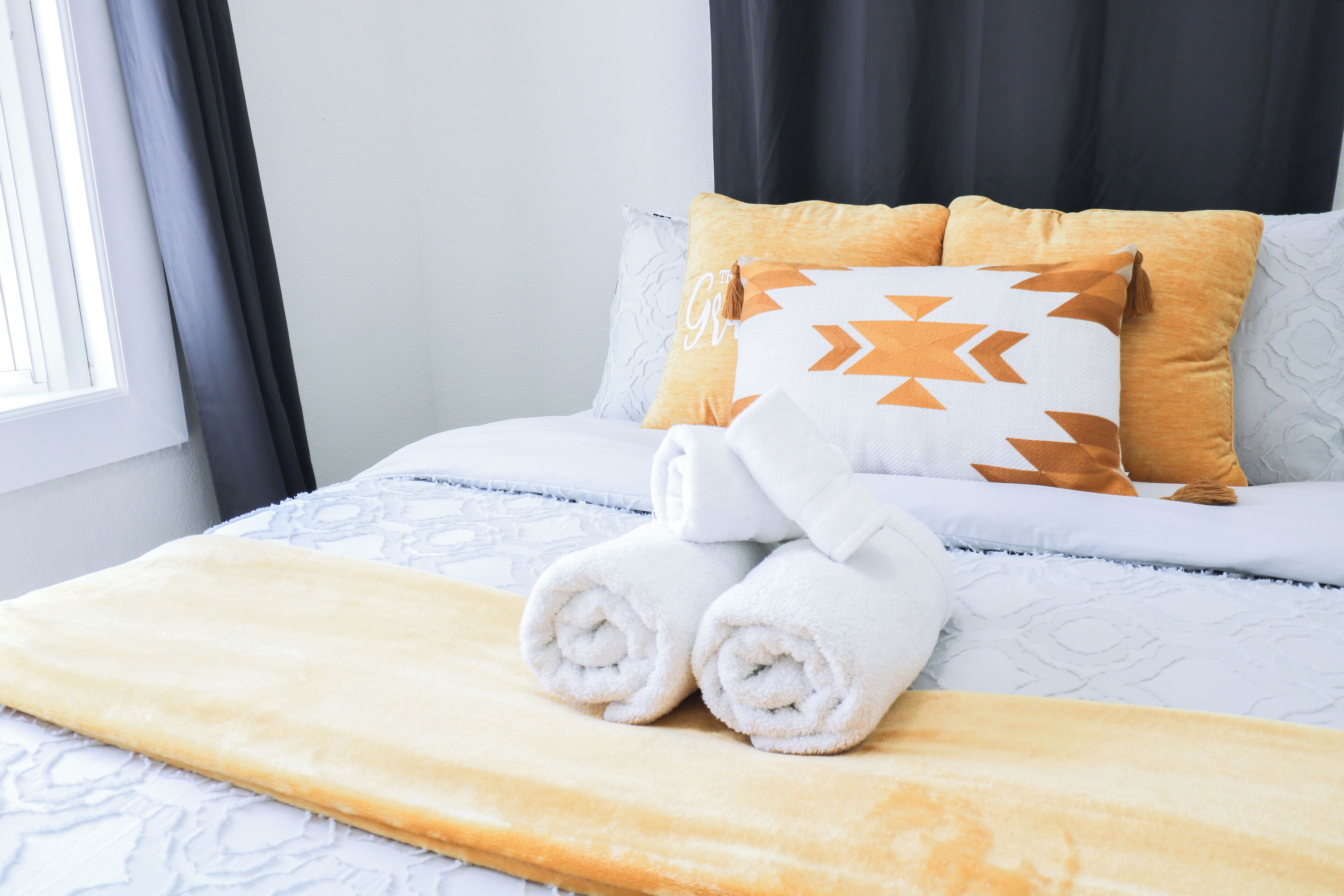 A neatly made bed with a white and orange patterned pillow, a rolled white towel, and a yellow blanket.