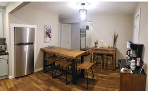 Una cocina compacta con refrigerador de acero inoxidable, una mesa de comedor de madera con bancos, un pequeño espacio para café y una puerta blanca.