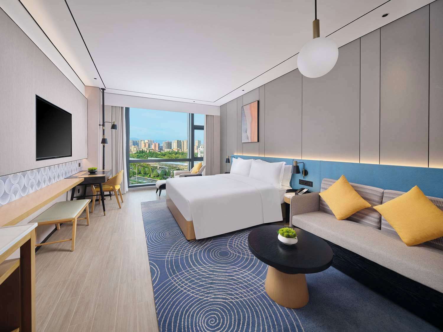 A modern hotel room with a large bed, a sofa, a desk, and a view of the city.