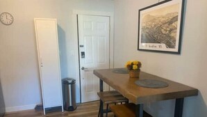 Un coin repas avec une table en bois, deux tabourets, une poubelle et un tableau encadré au mur.