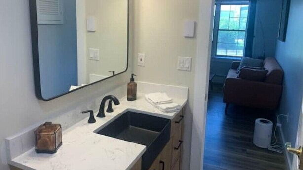 Une salle de bain avec un grand miroir, un lavabo et un plan de travail. Il y a une fenêtre avec des rideaux et une vue sur l’extérieur.