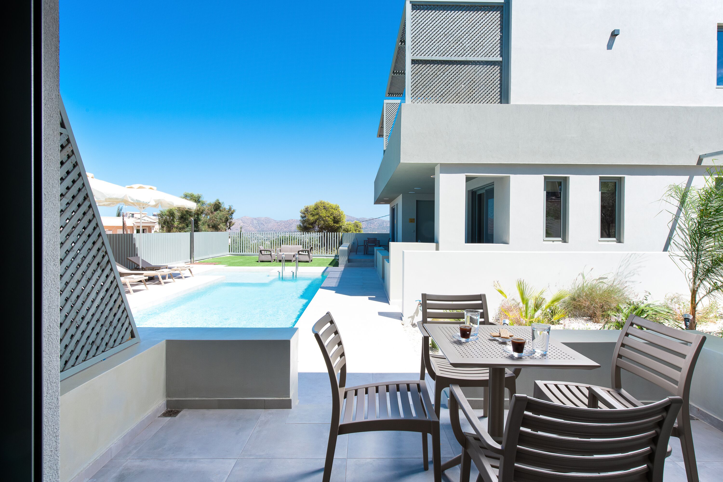 Ein moderner Poolbereich im Freien mit freiem Blick auf Berge, eine Terrasse mit Tisch und Stühlen sowie ein weißes Gebäude mit großen Fenstern.