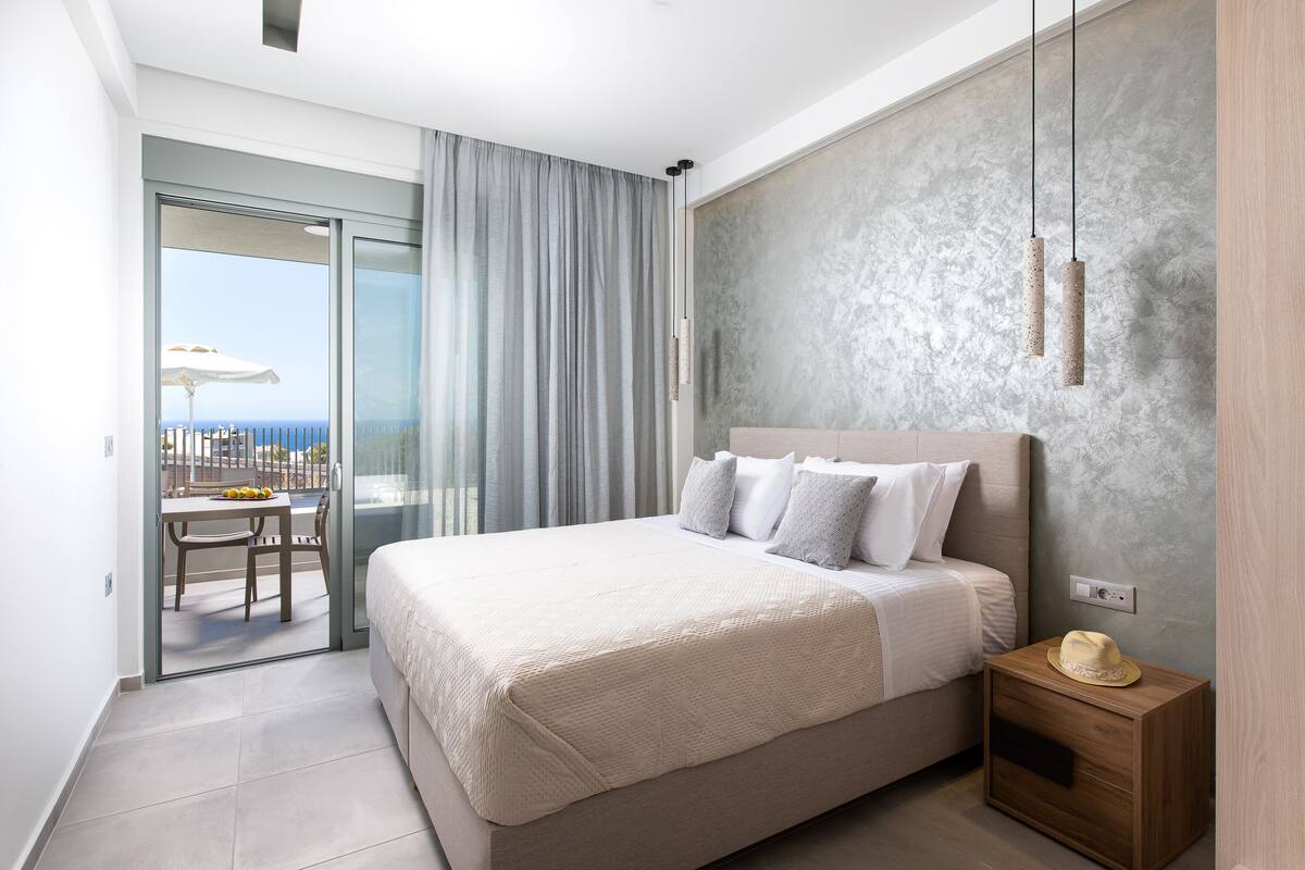 A bedroom with a bed, a nightstand, a sliding glass door to a balcony, and a view of the sea.