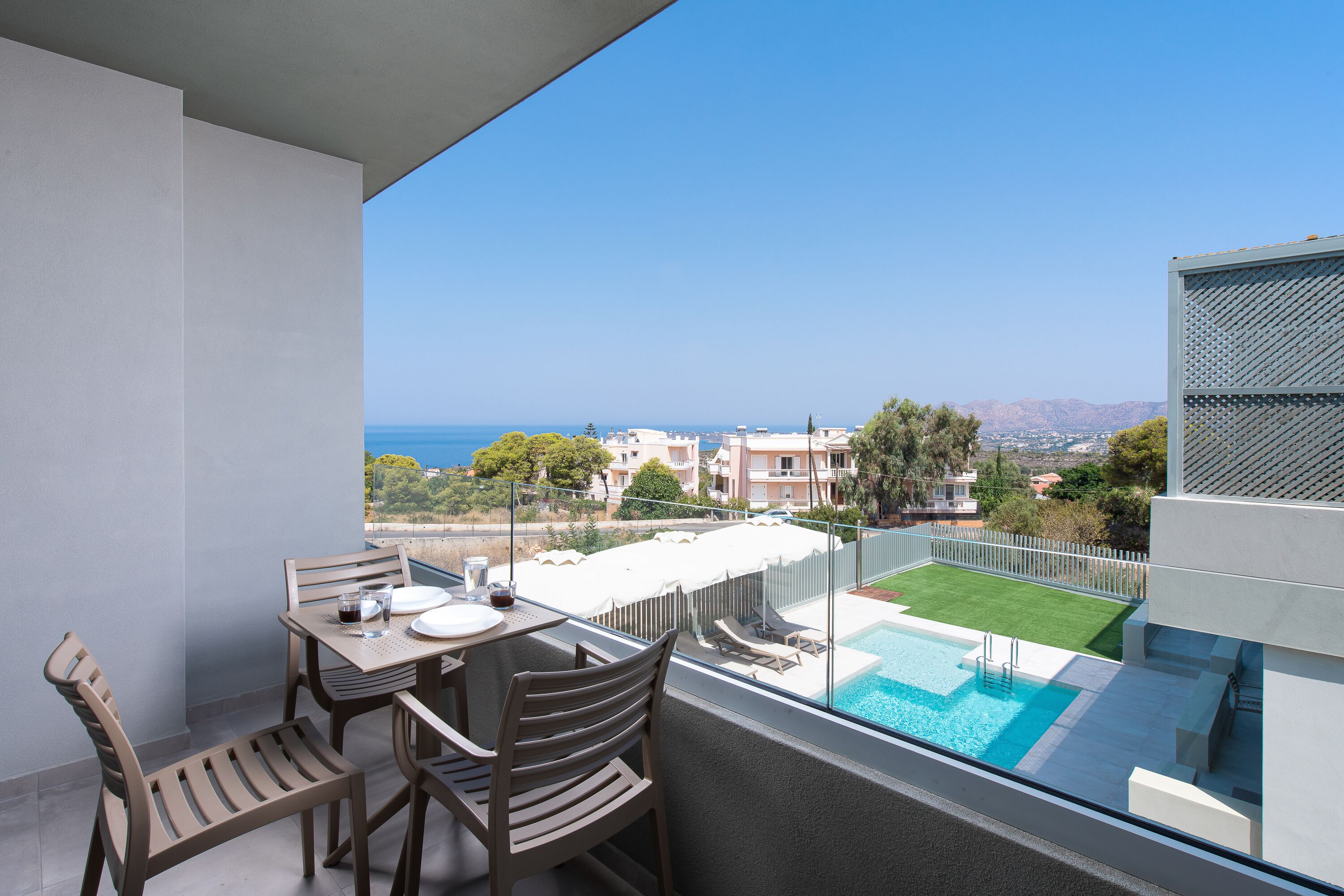 A balcony with a table and chairs, a swimming pool, and a view of the sea and buildings.