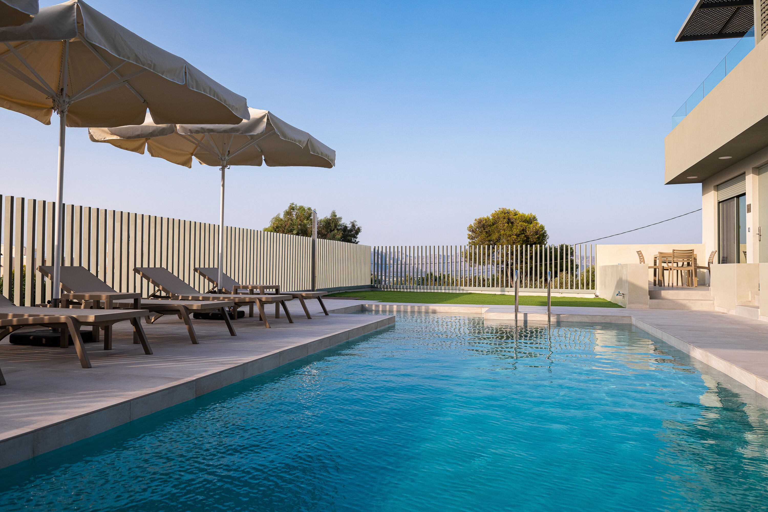 A swimming pool with lounge chairs and umbrellas, surrounded by a fence and a building.
