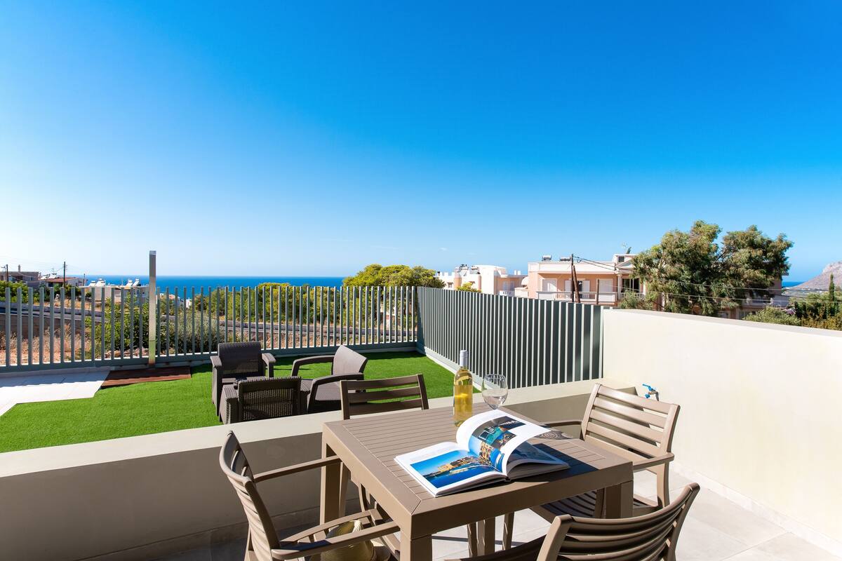 A terrace with a table, chairs, and a magazine. There is a view of the sea and a fence with greenery.
