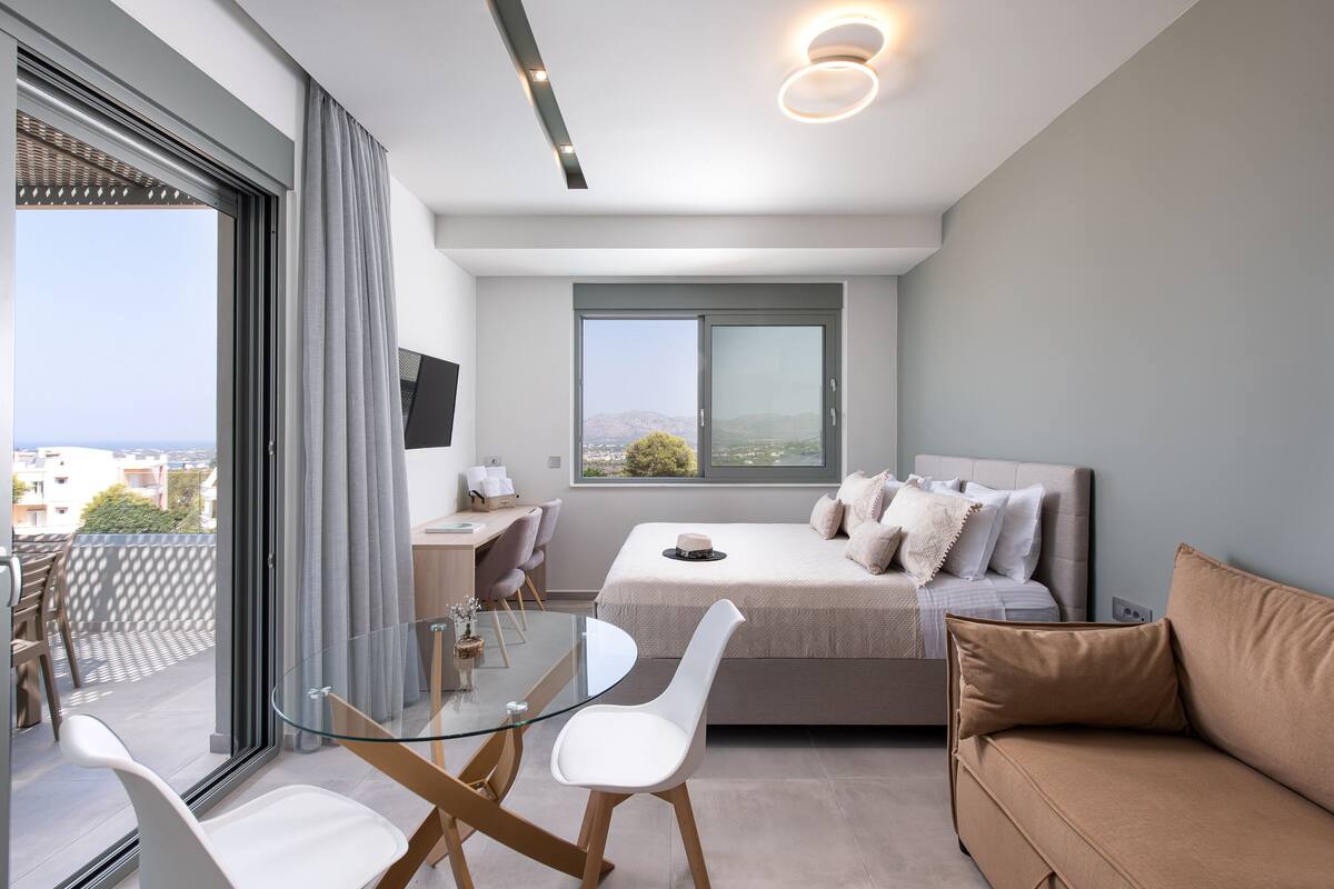 A modern bedroom with a glass dining table, a sofa, a bed with a view of the city, and a desk with a chair.