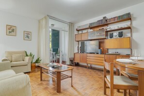 Una sala de estar moderna con un sofá, una mesa de comedor con sillas, una zona de cocina con armarios y electrodomésticos, y un ventanal corredero que da a un balcón.