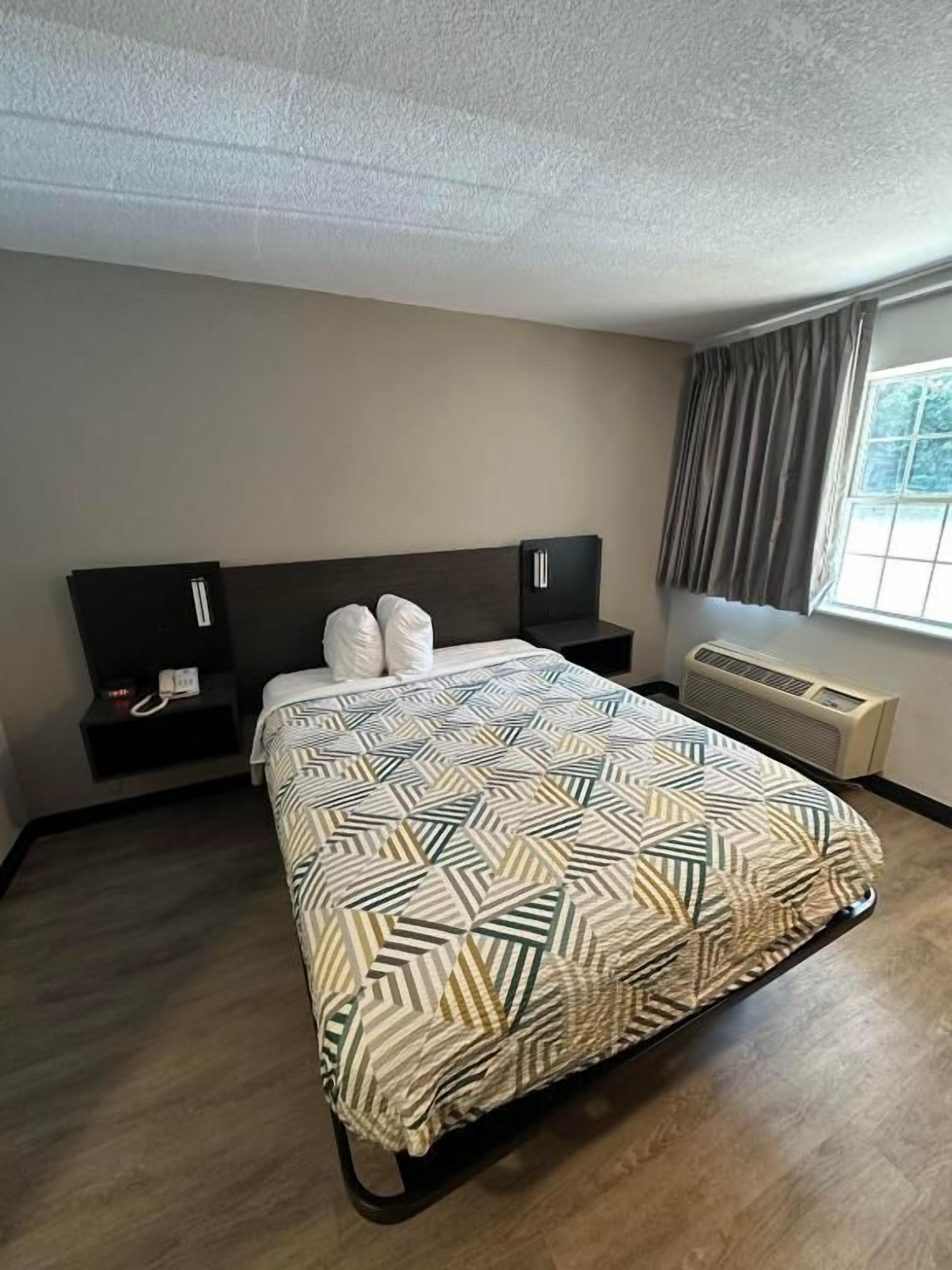 A hotel room with a bed, a bedside table, a heater, and a window with curtains.