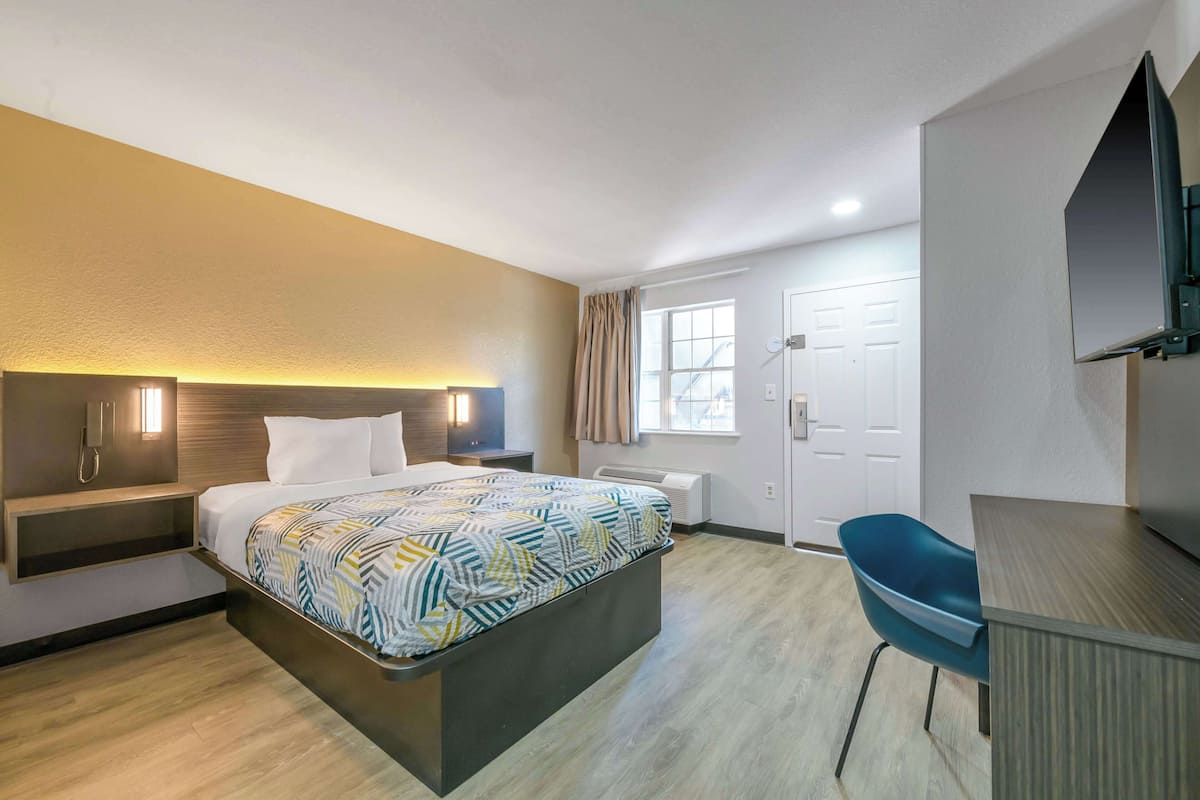 A modern hotel room with a bed, bedside table, lamp, and a desk with a chair.