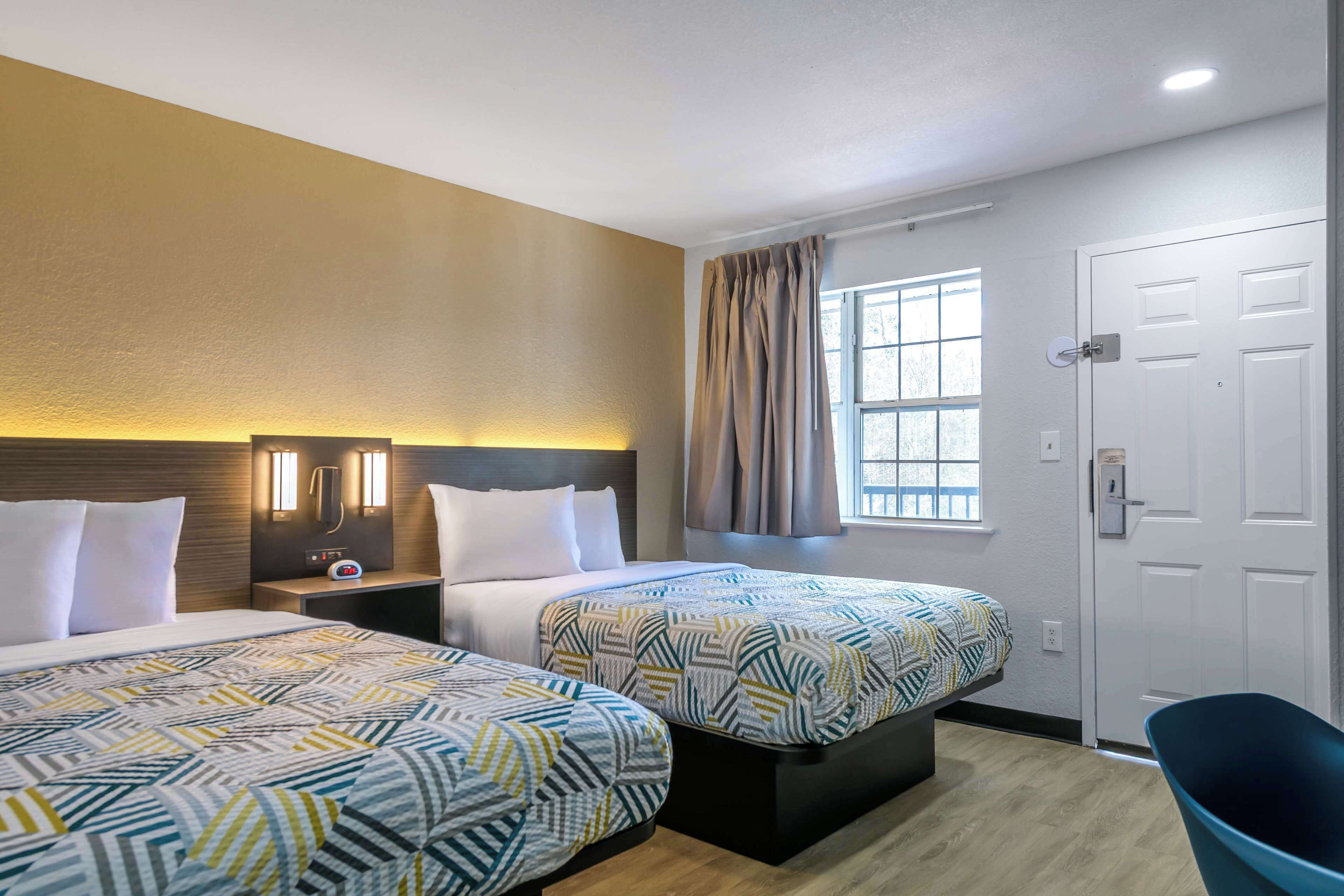 Une chambre d’hôtel avec deux lits, une table de chevet, une lampe fixée au mur, une fenêtre avec des rideaux et une porte.