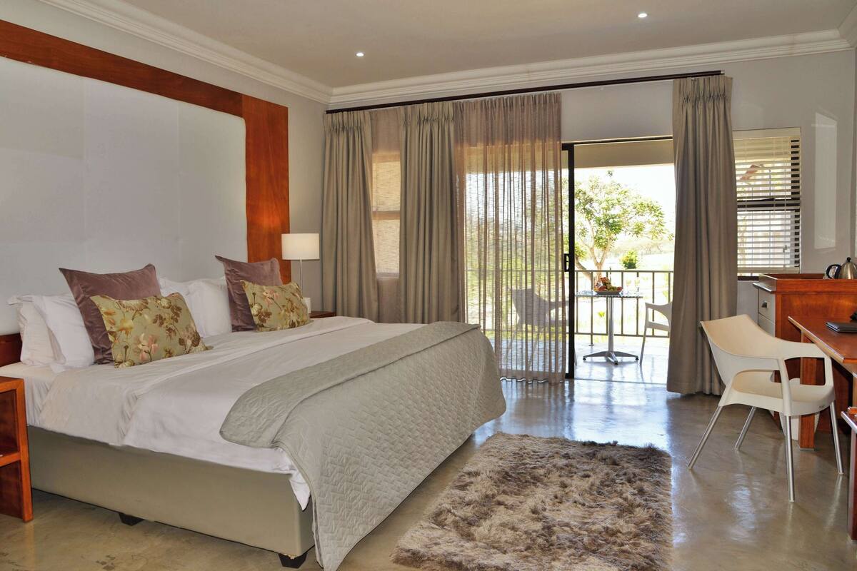 A bedroom with a large bed, two bedside tables, a desk with a chair, and a balcony with a table and chairs.