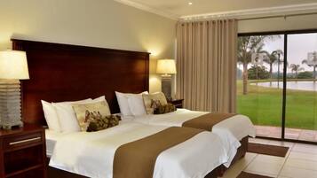Habitación de hotel con dos camas, cabecero de madera, dos mesitas de noche con lámparas y vista a un campo de golf a través de una puerta de vidrio.
