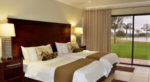 Une chambre d’hôtel avec deux lits, une tête de lit en bois, deux tables de chevet avec des lampes et une vue sur un terrain de golf, visible à travers une porte vitrée.