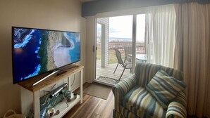 Een woonkamer met een televisie waarop een kustlandschap te zien is, een gestreepte fauteuil, en uitzicht op een terras met tuinmeubilair.