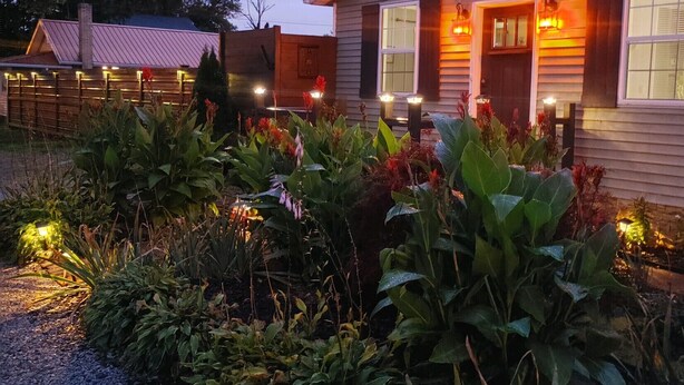 En stuga byggd av trä med en veranda, omgiven av en trädgård med olika växter och blommor.