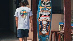 A person walking past a colorful surfboard with a tribal design, standing in front of a building with wooden pillars and a tiled floor. - Hospedaje Sueños DE Chicama, MAS Cerca AL Point, CON UNA Vista Expectacular (LA LIBERTAD)