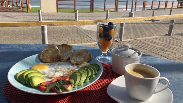 A table with breakfast items including toast, avocado, tomatoes, and a glass of fruit punch, with a view of the sea in the background. - Hospedaje Sueños DE Chicama, MAS Cerca AL Point, CON UNA Vista Expectacular (LA LIBERTAD)