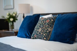 A neatly made bed with blue and floral pillows, a white bedspread, and a nightstand with a lamp and a vase of flowers. - Stonehenge Courtyard (Salisbury)