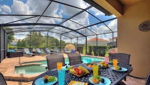 A covered outdoor dining area with a pool, lounge chairs, and a table set with food and drinks.