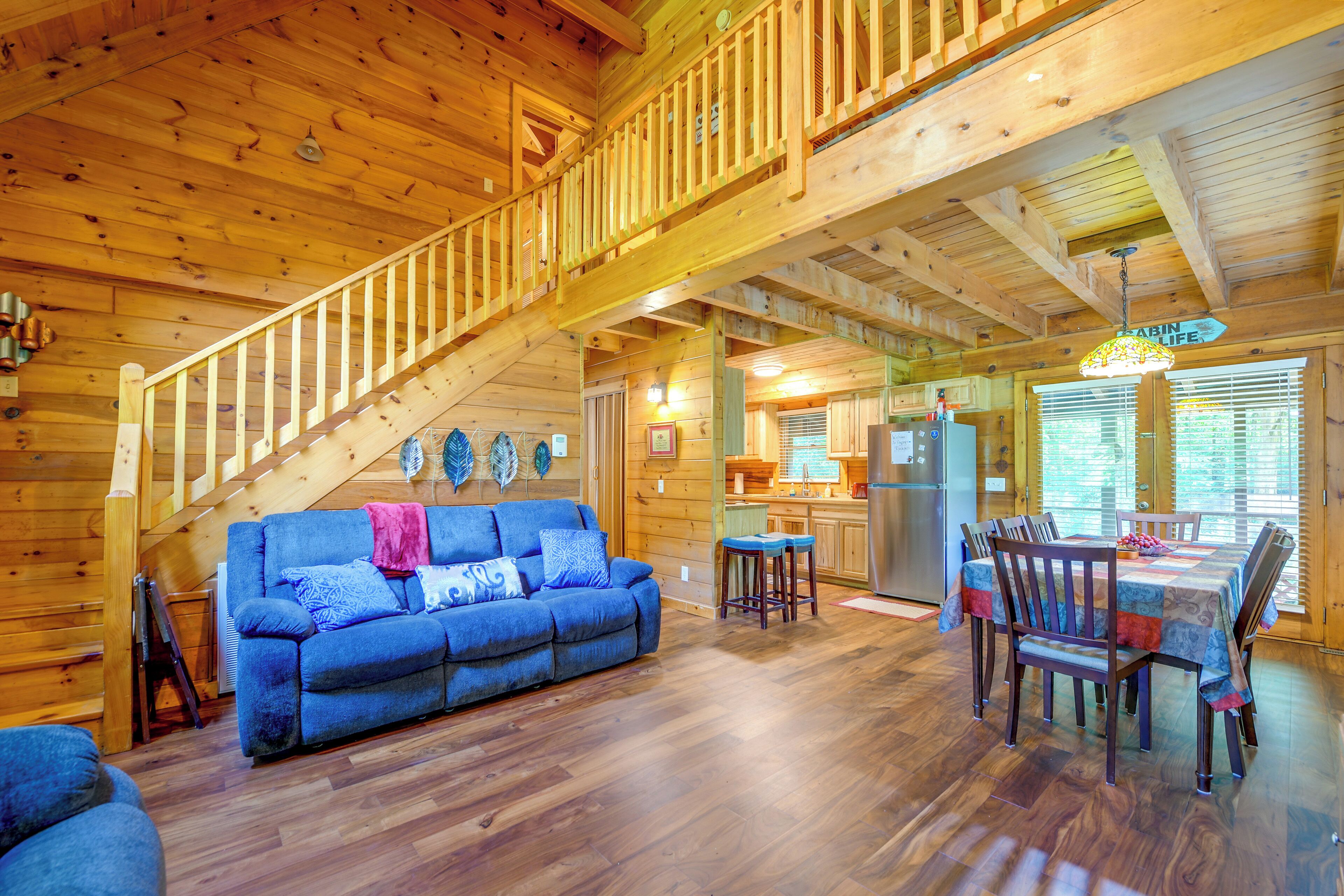 A cabin interior with a wooden staircase, a blue sofa, a dining table with chairs, and a kitchen area.
