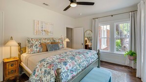 Un dormitorio con cama, ventilador de techo, ventana con cortinas y mesita de noche con lámpara.