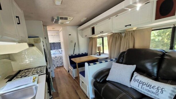 Interior compacto de uma RV, com cozinha compacta, área de refeições e espaço de estar com sofá de couro e almofada com a inscrição 'happiness is homemade'.