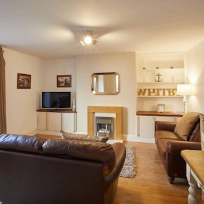 Een woonkamer met een bruine leren bank, een televisie, een open haard en een boekenplank met decoratieve objecten.