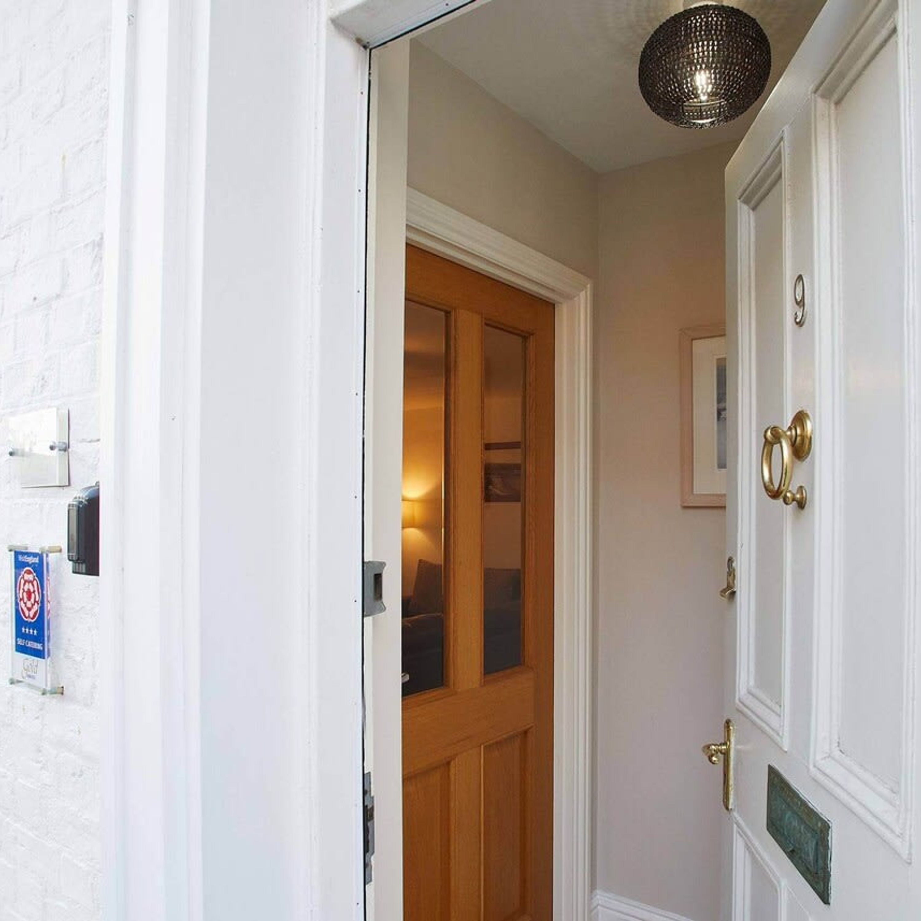 A white door with a brass handle and a number plate, a wooden door with glass panels, and a white wall with a doorbell and a sign.