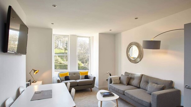 A modern living room with a sofa, a coffee table, a TV mounted on the wall, and large windows offering a view of the outdoors. - Host Stay Park View Apartment (Whitby)