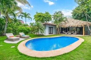 A swimming pool with lounge chairs, a small building with a thatched roof, and lush greenery.