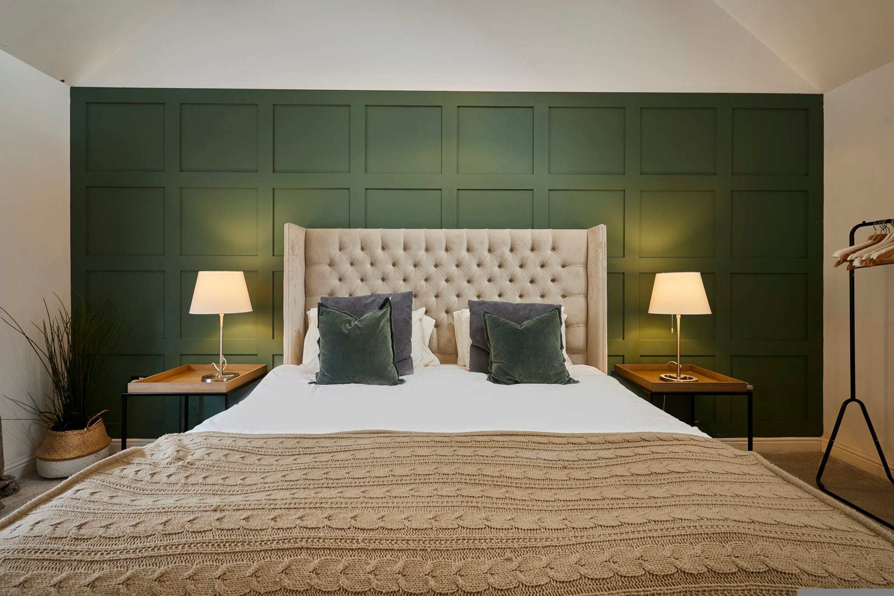 A bedroom with a tufted headboard, two bedside lamps, a large bed with a knitted blanket, and a green paneled wall.