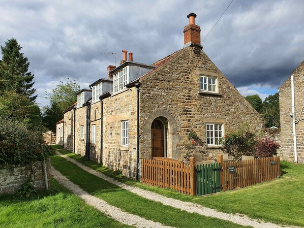 A stone building with a central arched doorway, multiple windows, and a gravel path leading to a wooden gate.
