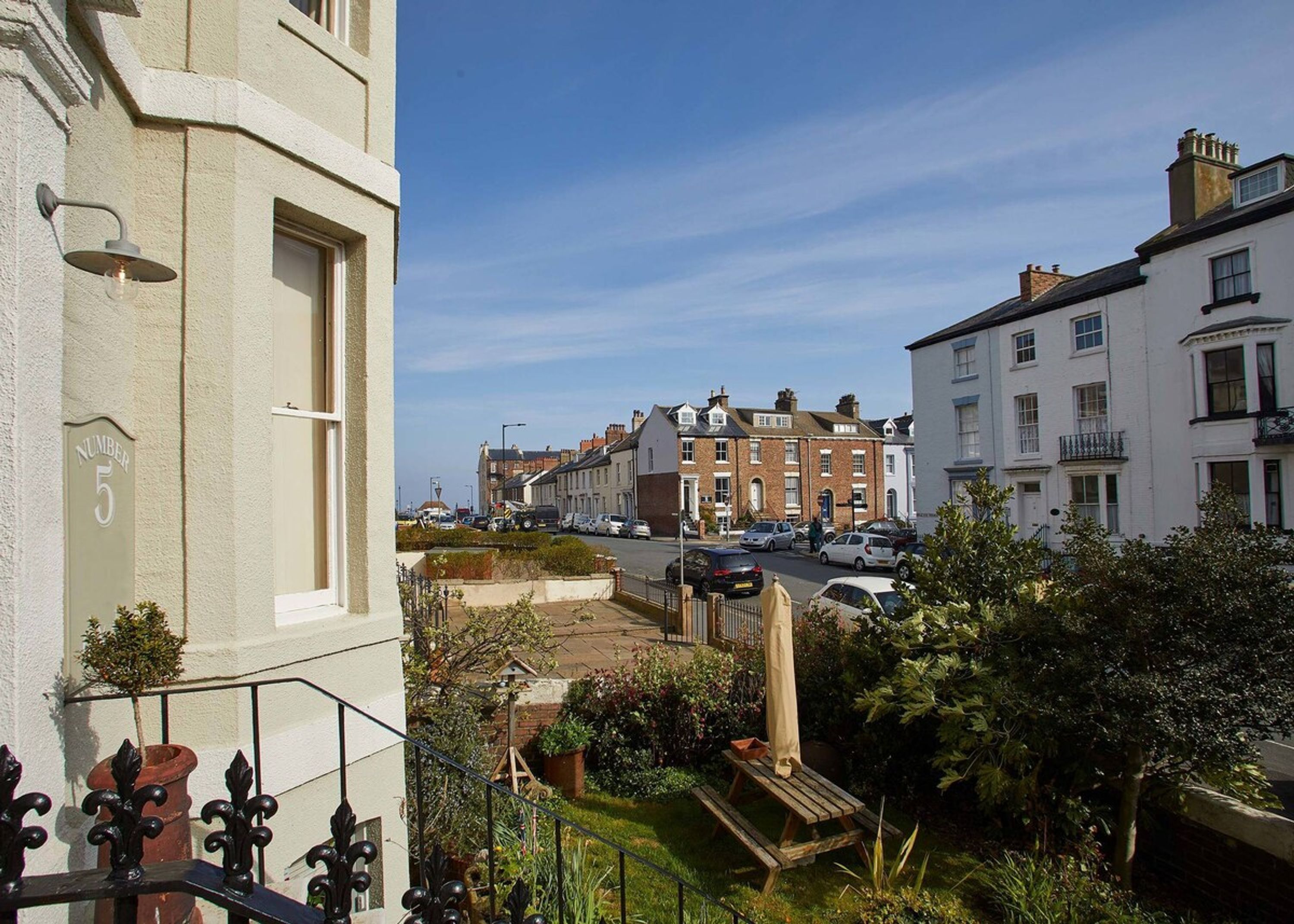 A residential street with a row of terraced houses, a clear sky, and a small garden area with a wooden bench.