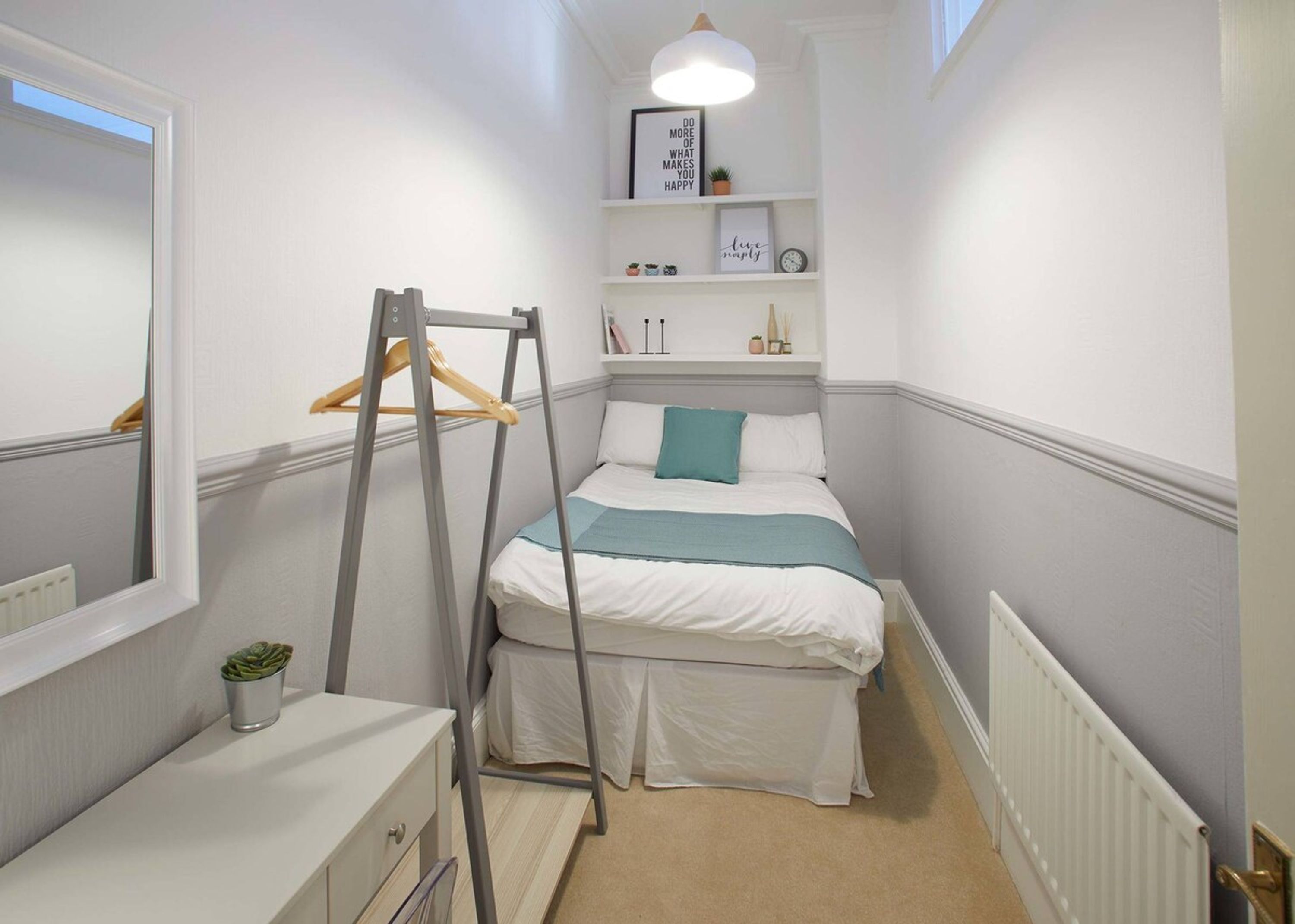 A narrow bedroom with a bed, a dresser, a mirror, a shelf with decorative items, and a ladder.
