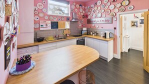 Une cuisine avec un mur rose, des armoires blanches et un plan de travail en bois. On peut y voir différents plats et ustensiles de cuisine, disposés sur le mur et sur le plan de travail.