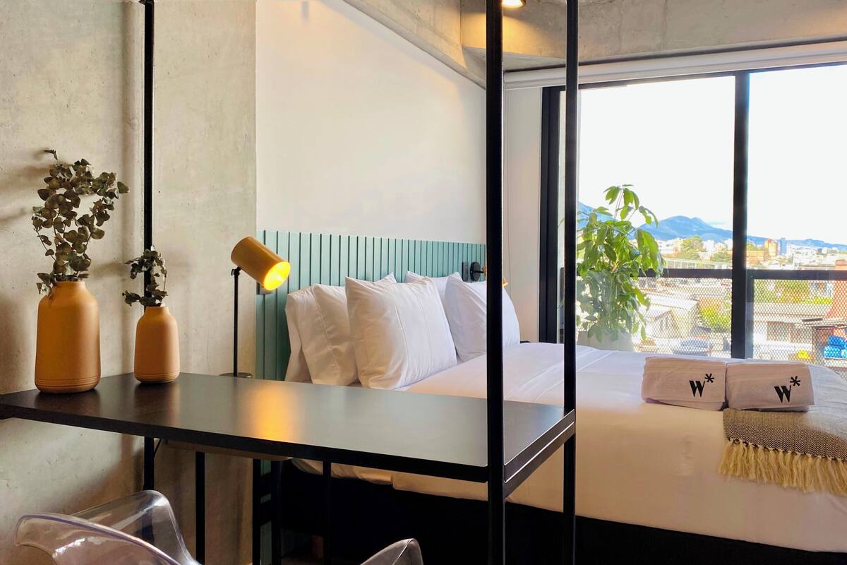 Une chambre d’hôtel moderne, équipée d’un lit, d’un bureau, de deux vases avec des plantes, d’une lampe et offrant une vue sur la ville grâce à de grandes fenêtres.
