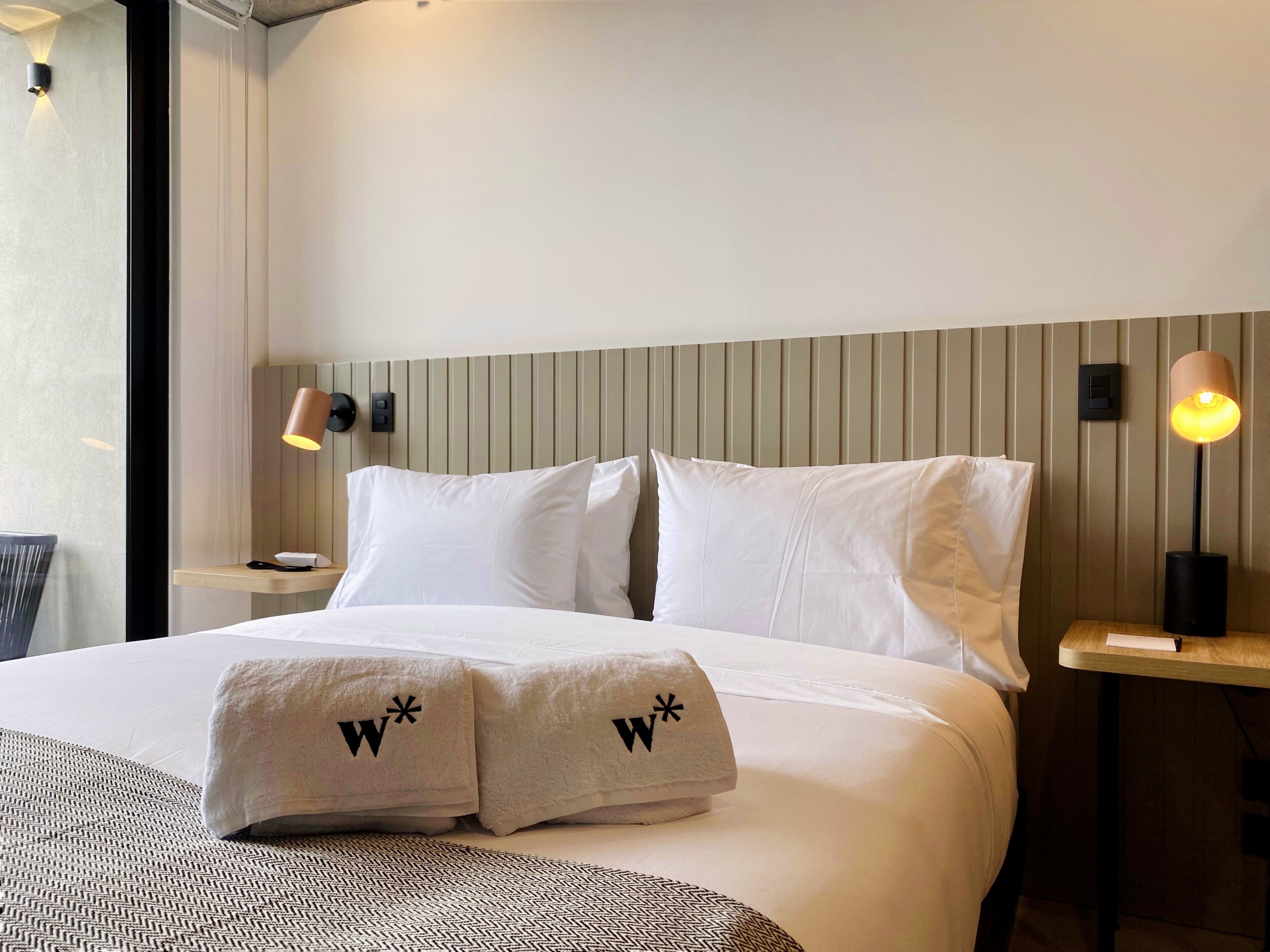 A hotel room with a neatly made bed, two pillows with a logo, a bedside table with a lamp, and a wall-mounted light fixture.