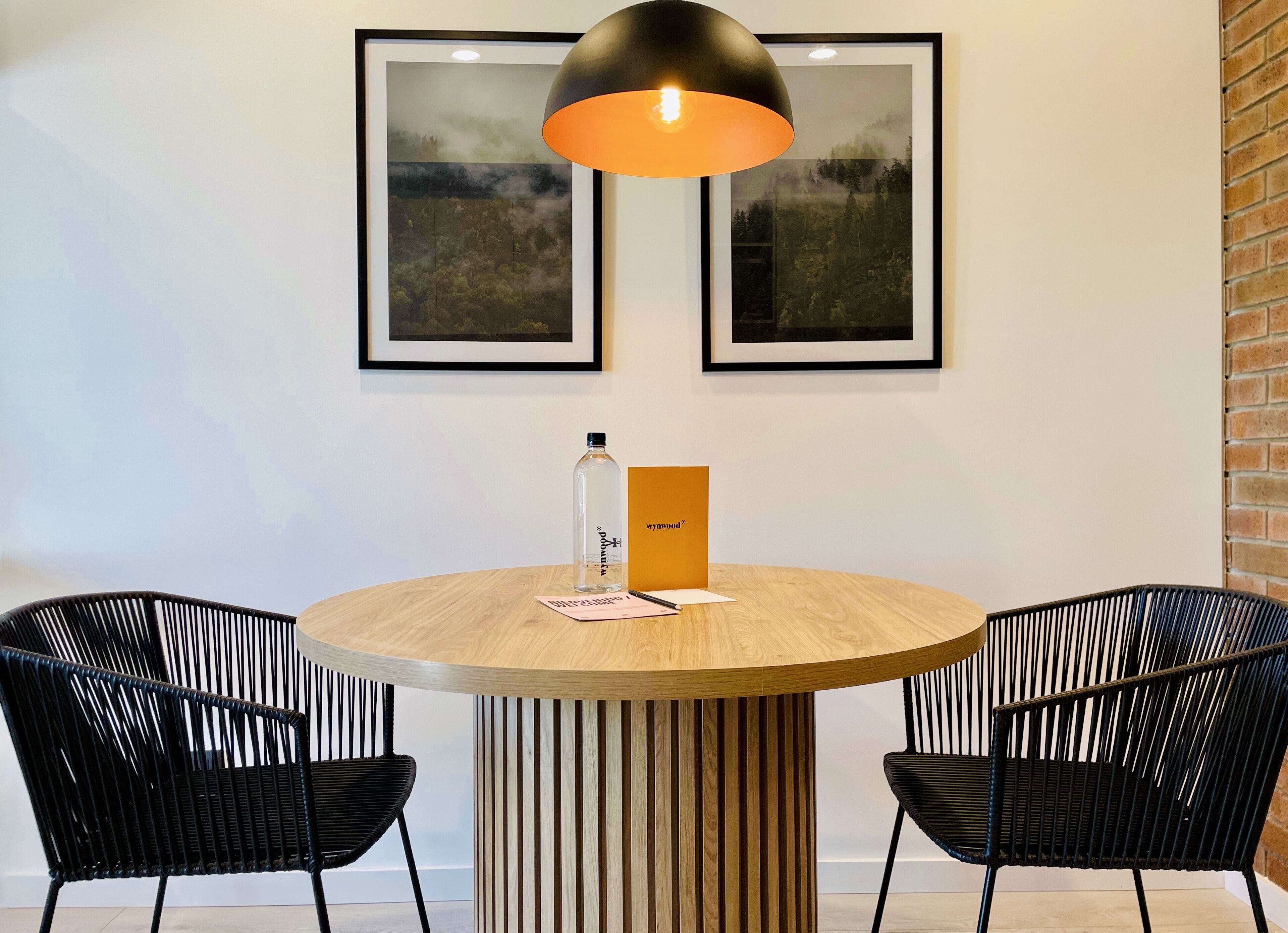 Une table ronde en bois, deux chaises noires, une bouteille d’eau, un menu et des tableaux encadrés accrochés au mur.