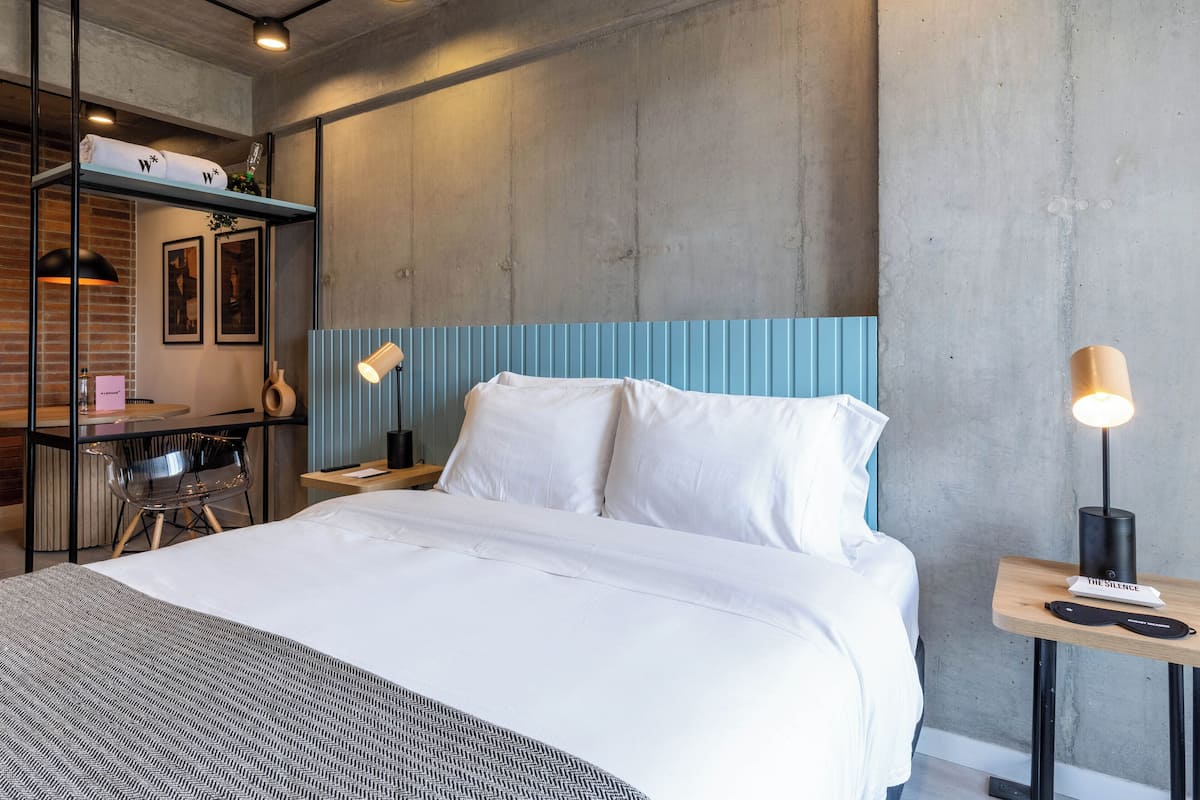Une chambre d’hôtel moderne équipée d’un lit, d’un bureau avec une chaise, d’une étagère avec des serviettes et de deux lampes fixées au mur.