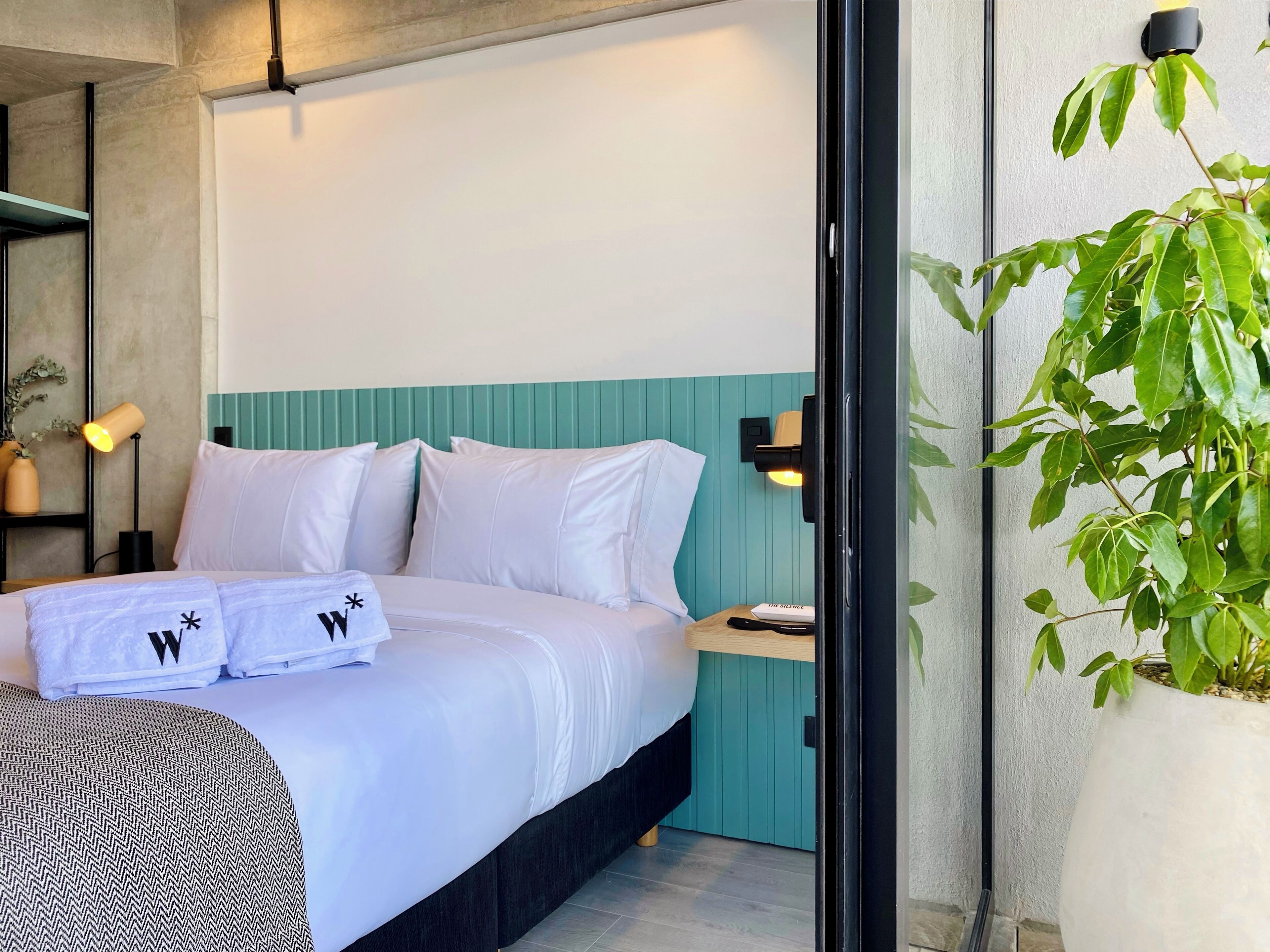 A neatly made bed with white linens and pillows, a bedside table with a book titled "The Silence," and a wall-mounted lamp.